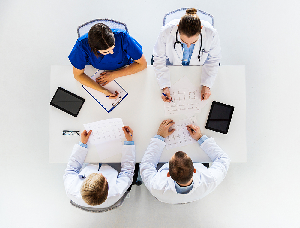 4 médicos sentados em uma mesa, 2 em cada lado, discutindo o que parece ser um exame cardíaco.